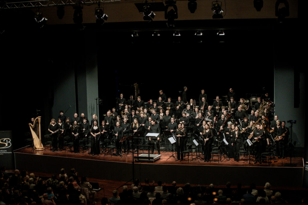 Orchester auf der Bühne mit vielen Blasinstrumenten, dunkler Saal, schwarzes Bühnenbild, Konzertatmosphäre, warmes Scheinwerferlicht