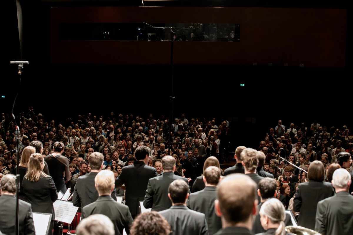 Konzertsaal mit großer Menschenmenge, dunkler Bühne und mehreren Musikern im dunklen Outfit mit Notenpulten, Blick auf Publikum und Orchester